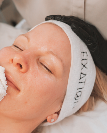 Person receiving a facial treatment with a white headband labeled 'FIXATION' on a neutral background.